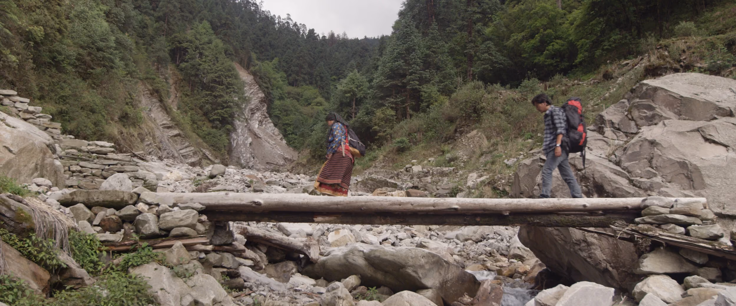 Two figures in wide scope framing against a Nepali hillside at dusk